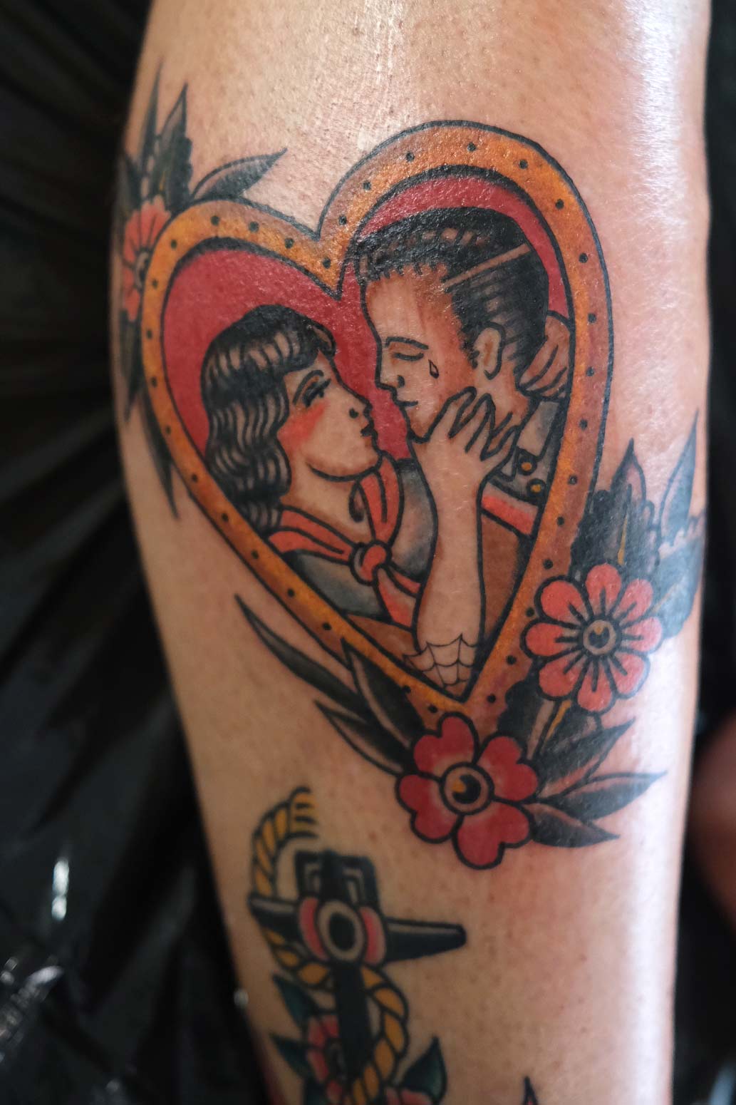 Oldschool Tattoo mit Herzmotiv, darin ein Liebespaar – umrankt von Blüten, gestochen im Tattoo Studio Good Old Times Berlin Mitte.